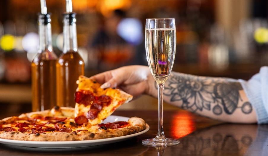 A hand lifting a slice of pizza from a plate with a glass of Prosecco in the foreground