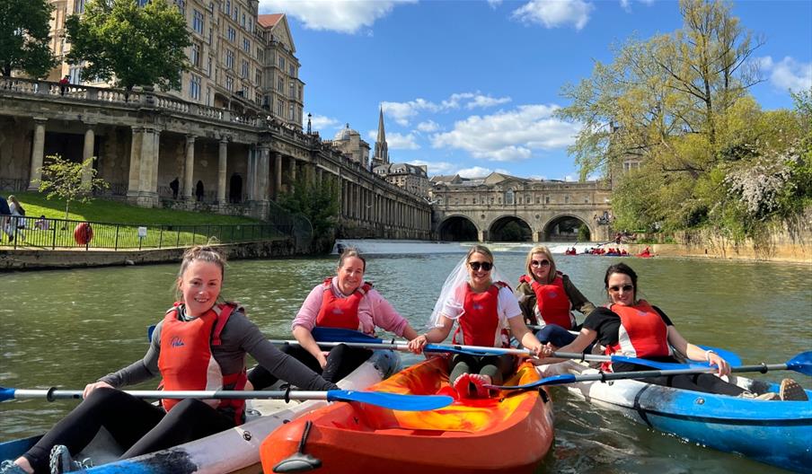 Photo of people, presumably on a bridal party given the attire, in kayaks in the river Avon near the Parade Gardens on a sunny day in Bath