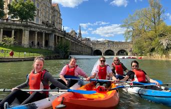 Photo of people, presumably on a bridal party given the attire, in kayaks in the river Avon near the Parade Gardens on a sunny day in Bath