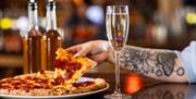 A hand lifting a slice of pizza from a plate with a glass of Prosecco in the foreground